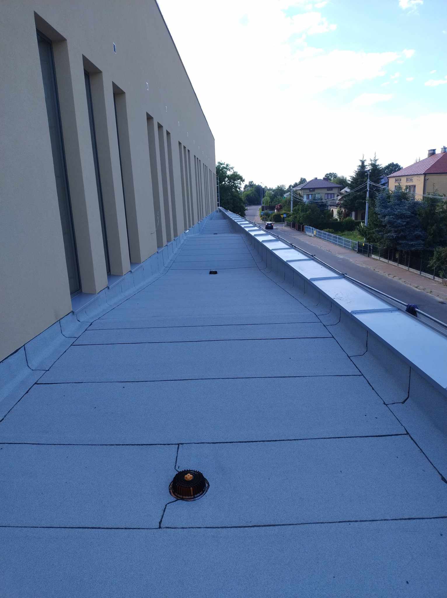 Widok z góry na dach płaski z szarą membraną, z widoczną obróbką blacharską i elementami wentylacyjnymi. W tle ulica i budynki.