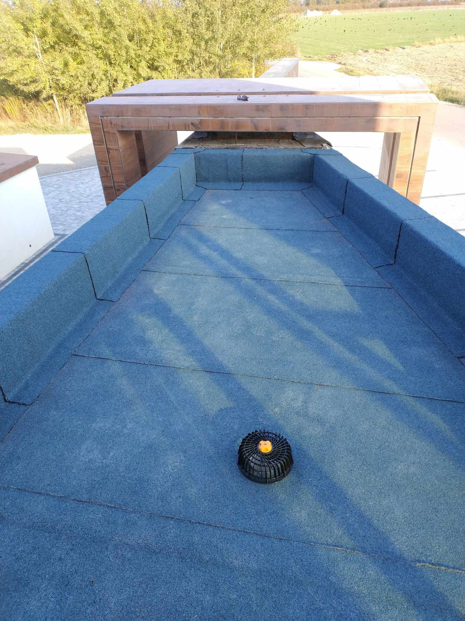 Widok z góry na część dachu płaskiego z membraną, z widocznym elementem wentylacyjnym i drewnianą konstrukcją w tle.