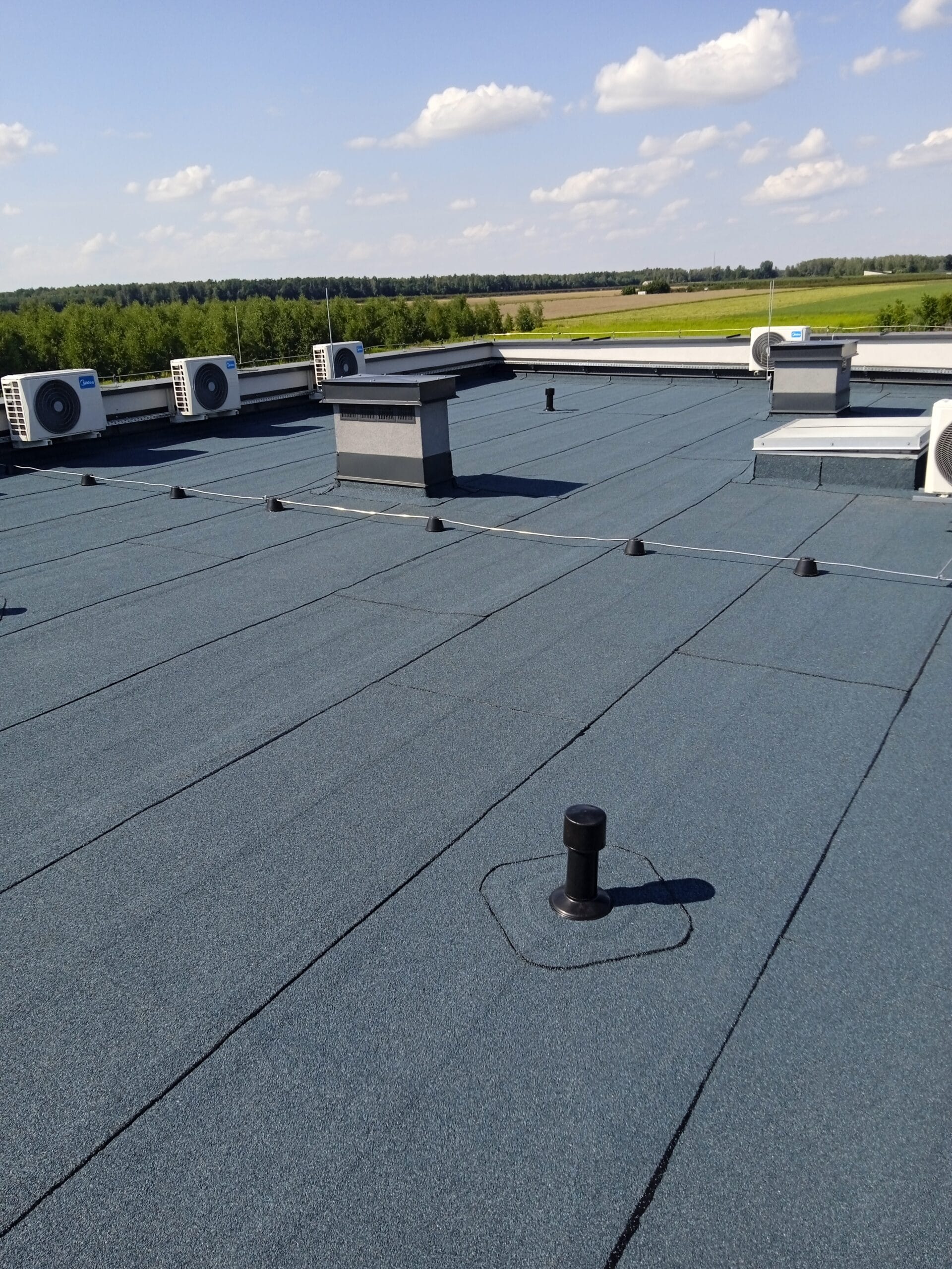 Szczegółowe zdjęcie dachu płaskiego z membraną, na którym znajdują się klimatyzatory, kominy wentylacyjne i punkty mocowania instalacji. Dach w kolorze ciemnoszarym, tło z zielenią.